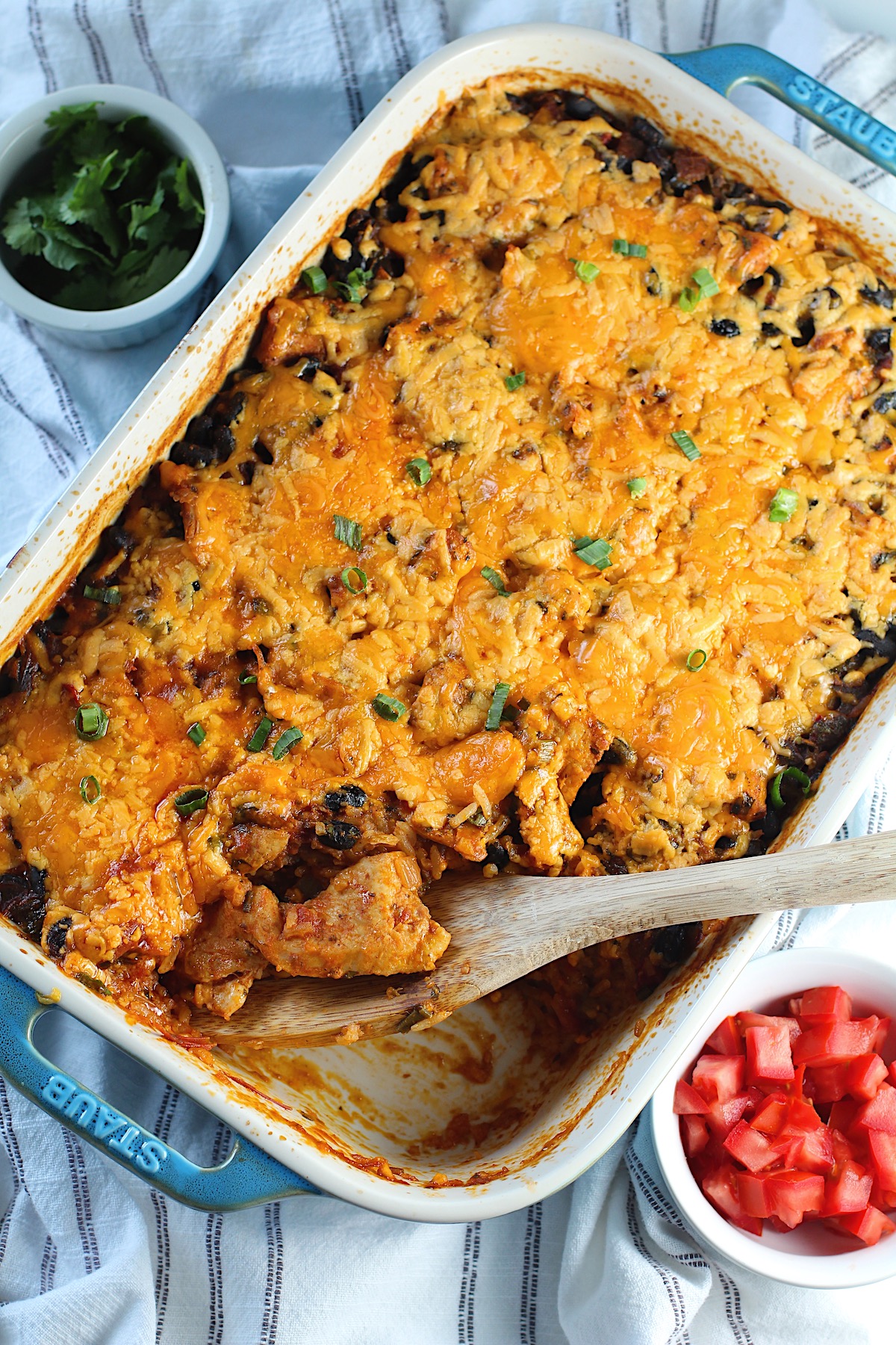 Salsa Chicken Casserole Recipe with cheese melted on top and scallions sprinkled over. A spatula is sitting in the corner where some of the casserole has been removed. A bowl of tomatoes is in the lower right and cilantro in the upper left.