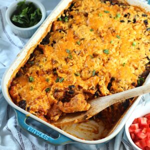 Salsa Chicken Casserole Recipe with cheese melted on top and scallions sprinkled over. A spatula is sitting in the corner where some of the casserole has been removed. A bowl of tomatoes is in the lower right and cilantro in the upper left.