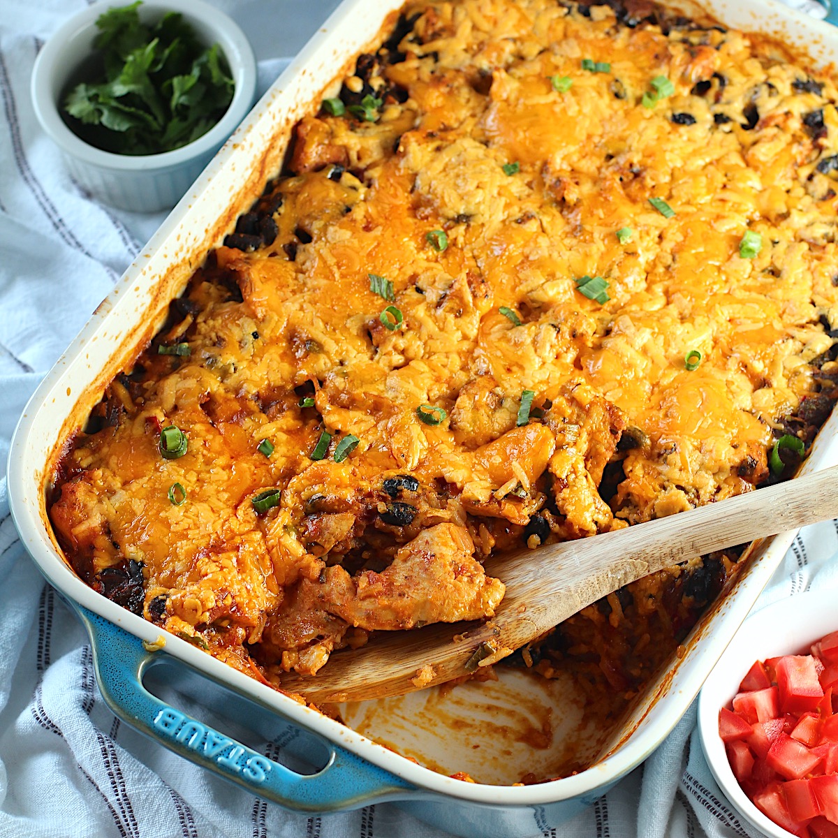Salsa Chicken Casserole Recipe with cheese melted on top and scallions sprinkled over. A spatula is sitting in the corner where some of the casserole has been removed. A bowl of tomatoes is in the lower right and cilantro in the upper left.