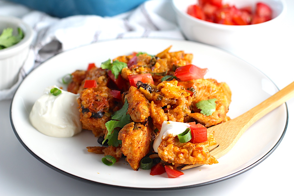 Salsa Chicken Casserole Recipe with rice on a plate with bamboo fork, dollop of sour cream on the side, and cilantro leaves on top. In the background, the casserole dish, a small bowl with diced fresh tomatoes and another bowl with cilantro leaves..
