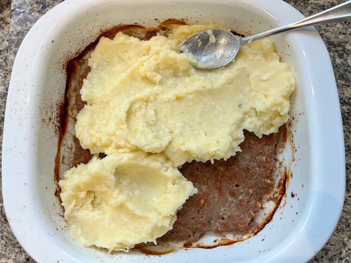 Cooked meatloaf in the bottom of a casserole pan and spoon spreading mashed potatoes over top for Meatloaf Casserole Recipe.