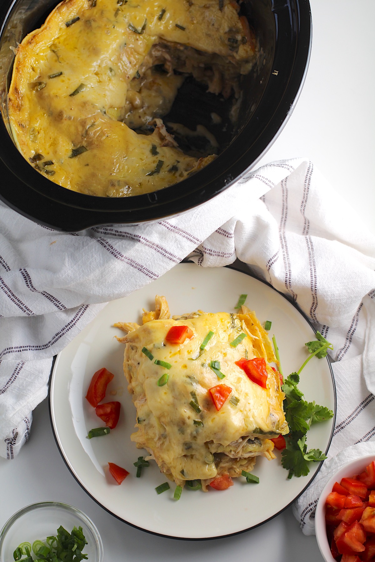 Piece of layered Crockpot White Chicken Enchilada Casserole on a plate with diced tomato, sliced scallion, and cilantro garnish. Crockpot with casserole is in the background and kitchen towel behind plate.