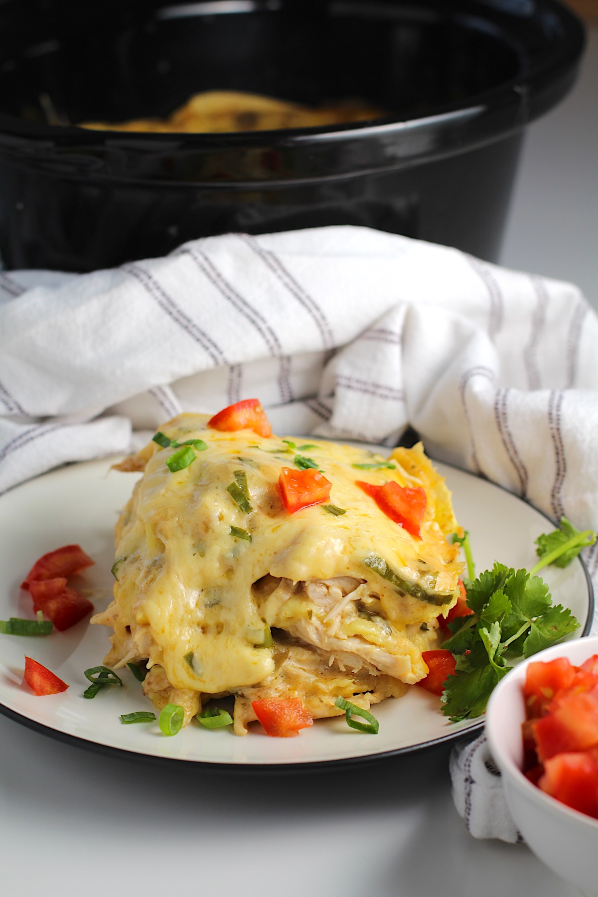 Piece of layered Crockpot White Chicken Enchilada Casserole on a plate with diced tomato, sliced scallion, and cilantro garnish.  Crockpot with casserole is in the background and kitchen towel behind plate.