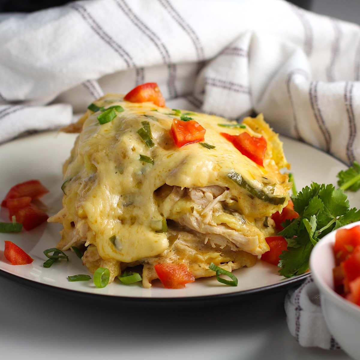 Piece of layered Crockpot White Chicken Enchilada Casserole on a plate with diced tomato, sliced scallion, and cilantro garnish. Kitchen towel behind plate.