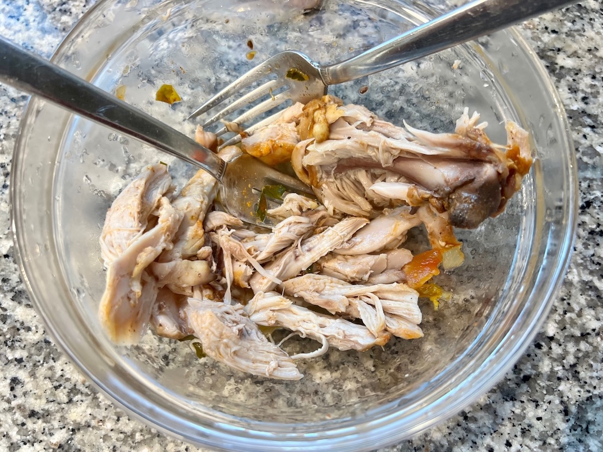 Forks pulling and shredding chicken thigh meat from the bones in a glass bowl for Spanish Chicken Soup Recipe.