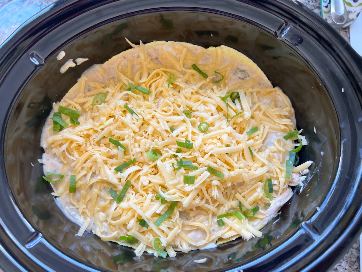 All layers added to the slow cooker insert with shredded cheese and scallion slices on top before cooking for crockpot white chicken enchilada casserole.