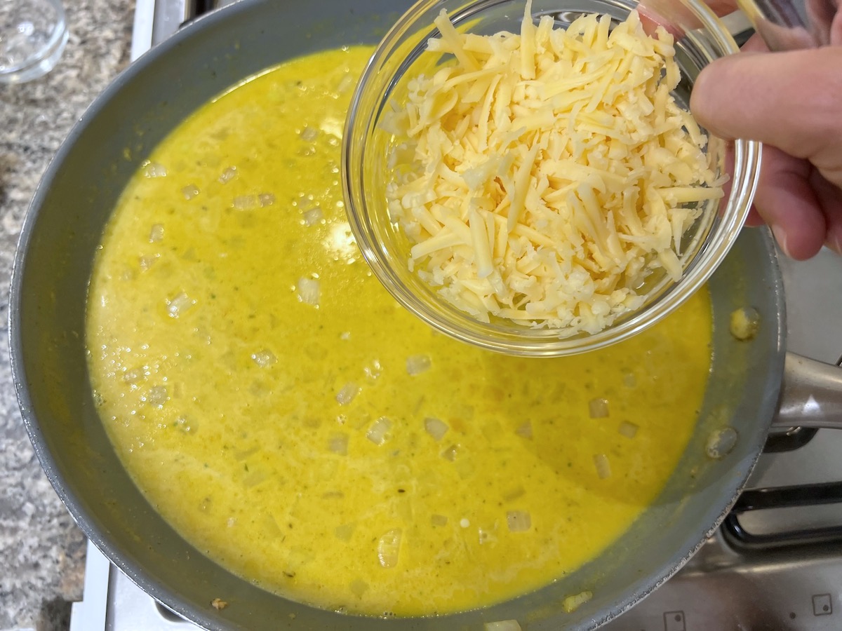 Hand holding a bowl of shredded white cheddar over a frying pan with the curry sauce for Chicken Divan Curry Casserole Recipe.