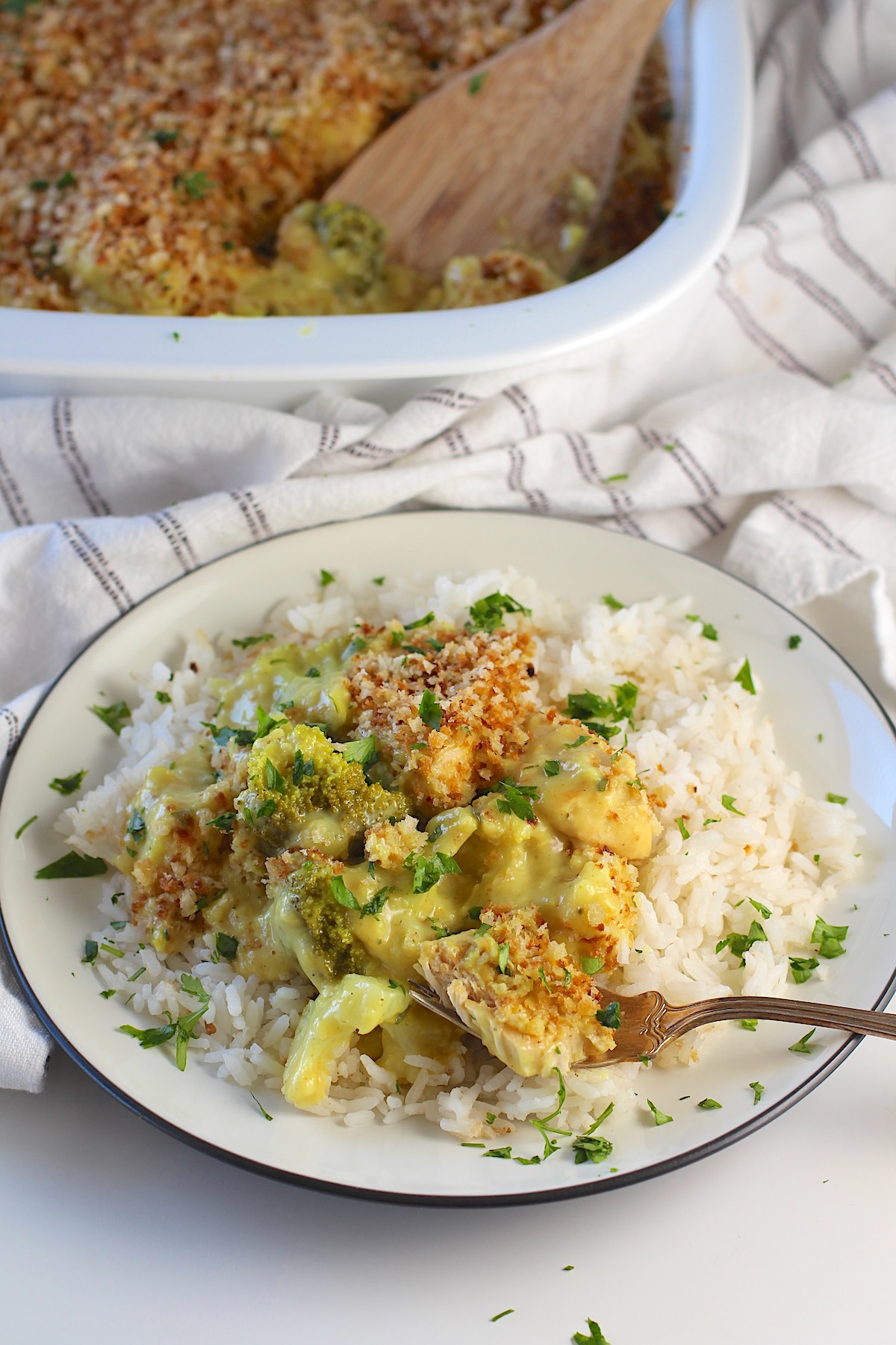 Creamy Broccoli Chicken Curry Casserole Recipe served over white rice on a plate with fork scooping up a bite. Plate is on a counter with dish towel and casserole dish behind it.