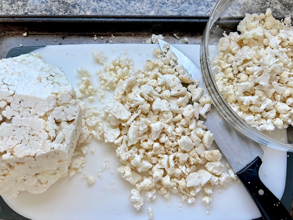 Cauliflower chopped into small pieces (cauliflower rice) on a cutting board with knife and bowl of cauliflower rice next to it for Low carb Cauliflower Rice and Chicken Casserole.