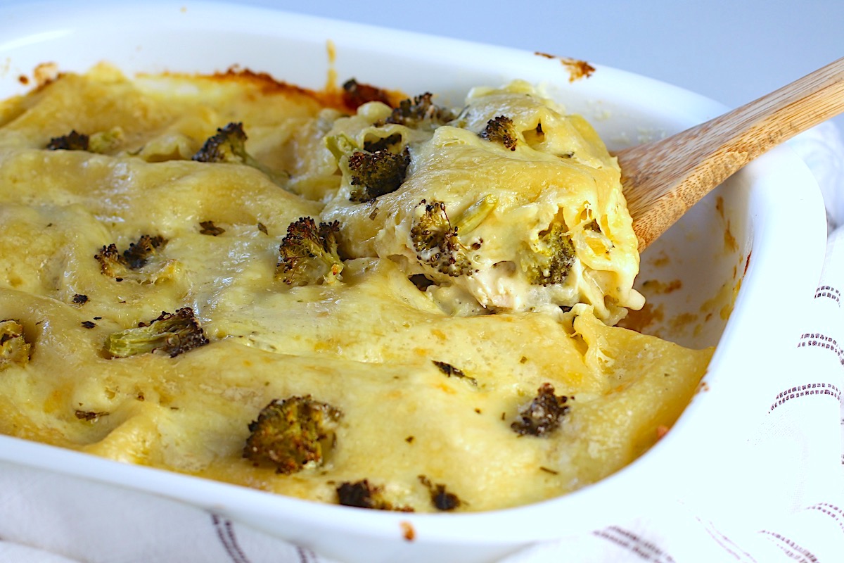 Broccoli and Chicken Lasagna in casserole dish with spatula scooping up a piece from the corner showing creamy and cheesy filling.