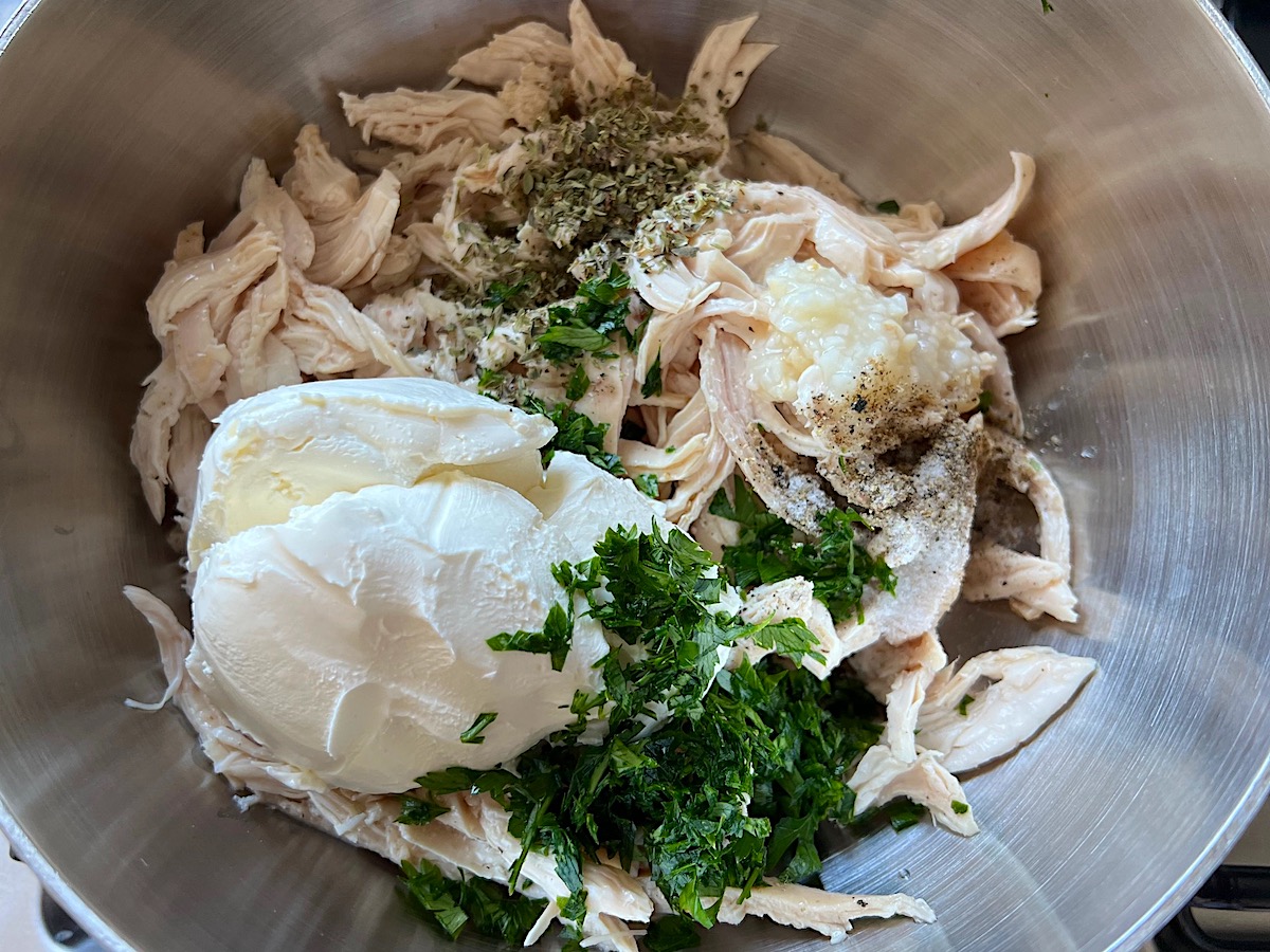 Cooked shredded chicken, softened cream cheese, parsley, garlic, oregano, salt, and pepper in a metal bowl for Broccoli and Chicken Lasagna recipe.
