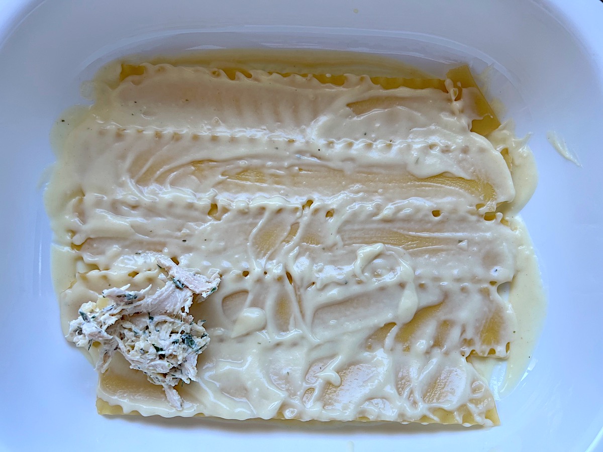 Lasagna noodles covered in bechamel sauce layered at the bottom of a casserole dish with a shredded chicken and cheese filling scoop added to one corner for Broccoli and Chicken Lasagna Recipe.