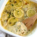 Creamy Chicken Piccata Casserole Recipe in a casserole dish with lemon slices and parsley on top. A wooden spatula is scooping a piece of chicken in the creamy sauce up from the corner.
