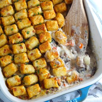Spatula scooping the corner of a creamy and cheesy hamburger tater tot casserole with golden brown tater tots on top all lined up in the casserole dish on the counter.