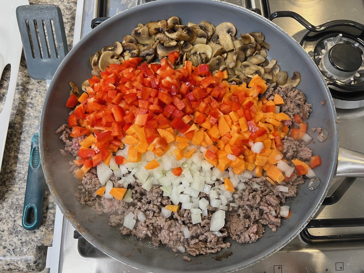 Mushroom slices, diced red pepper, diced carrots, diced onion, and ground beef in large pan cooking on stove for Cheesy Hamburger Tater Tot Casserole Recipe.
