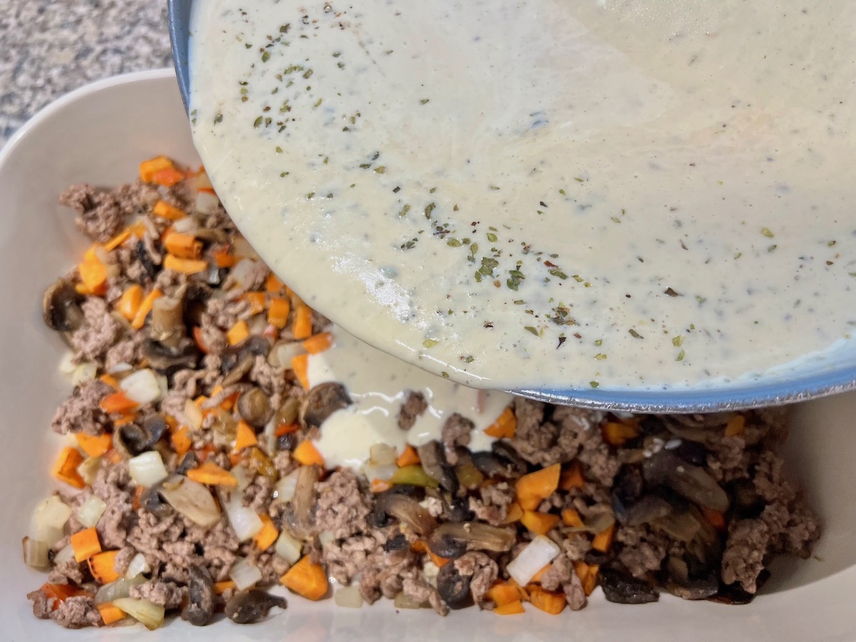 Pan pouring warm thickened sauce over ground beef and veggies in casserole dish for Cheesy Hamburger Tater Tot Casserole Recipe.