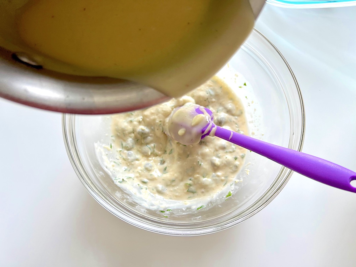 Pot pouring warm thickened broth, butter, flour sauce into mayonnaise and caper sauce in a clear mixing bowl with spatula for the Creamy Chicken Piccata Casserole recipe.