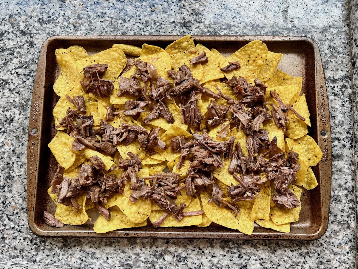 Shredded Beef layered on top of yellow corn tortilla chips on a large sheet pan on counter for Birria Nachos.