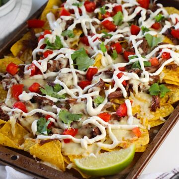 Birria Nachos on a sheet pan with yellow corn toritlla chips topped with tender shredded beef, melty cheese, a drizzle sour cream, fresh diced tomatoes, cilantro leaves, and a lime wedge in the corner of the pan.