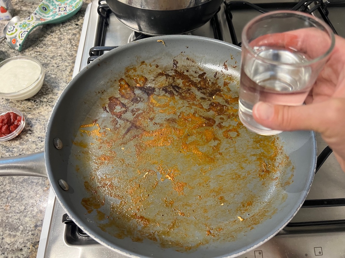 Hand holding a small measuring cup of vodka over the pan where the chicken was cooked for the Chicken and Vodka pasta recipe.