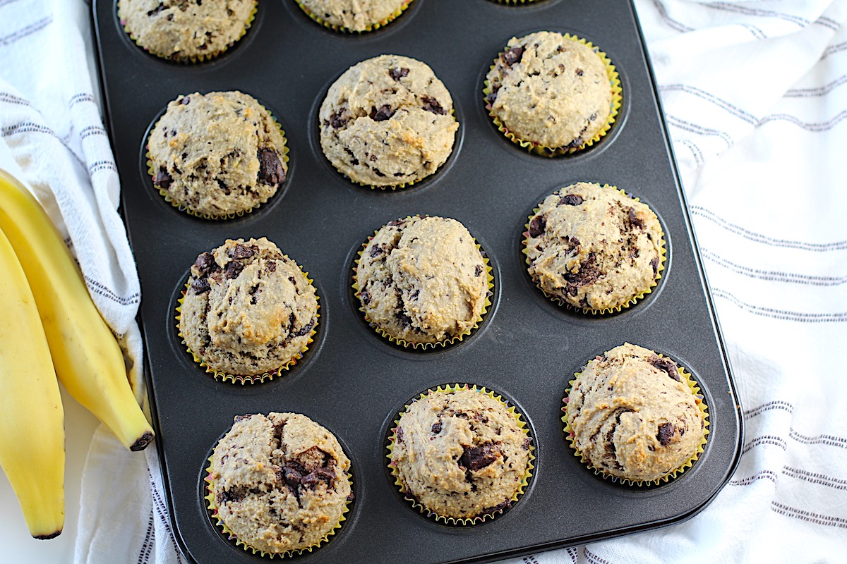 Oat Banana chocolate chip muffins freshly baked in a muffin tin with fresh bananas laying beside it.