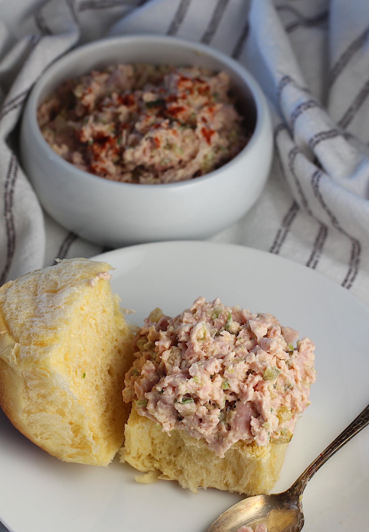 Old fashioned ham salad topping an open face bun, served on a plate with a spoon resting beside it. 