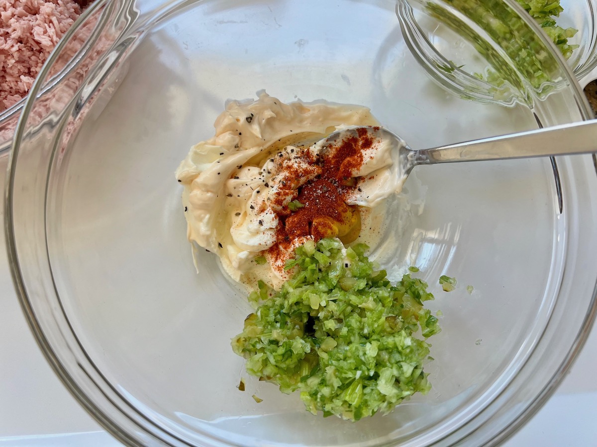 Seasonings, mayonnaise and pickle relish measured int a glass mixing bowl with a spoon resting into it.