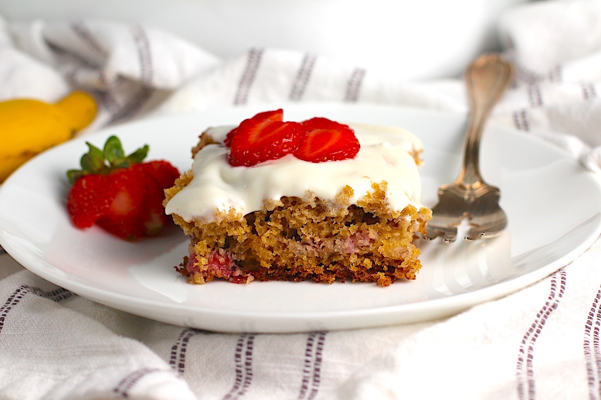 Piece of Strawberry Banana Cake with Vanilla Icing on a plate with a fork with sliced strawberries on top and a strawberry sliced and fanned out to the left.