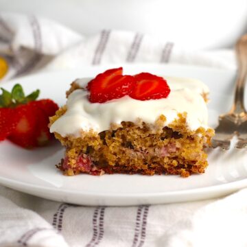Piece of Strawberry Banana Cake with Vanilla Icing on a plate with a fork with sliced strawberries on top and a strawberry sliced and fanned out to the left.