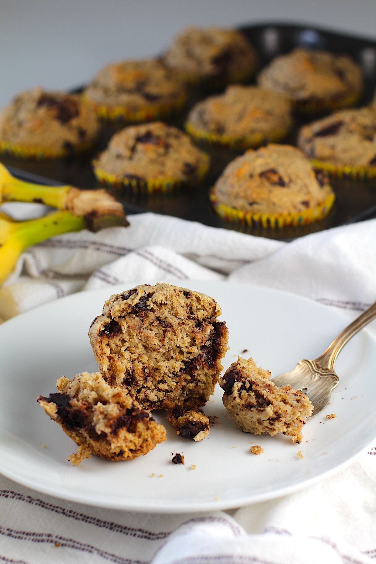 An oat banana chocolate chip muffin on a plate with pieces of the muffin cut away and one on a fork, that shows the melted chocolate.