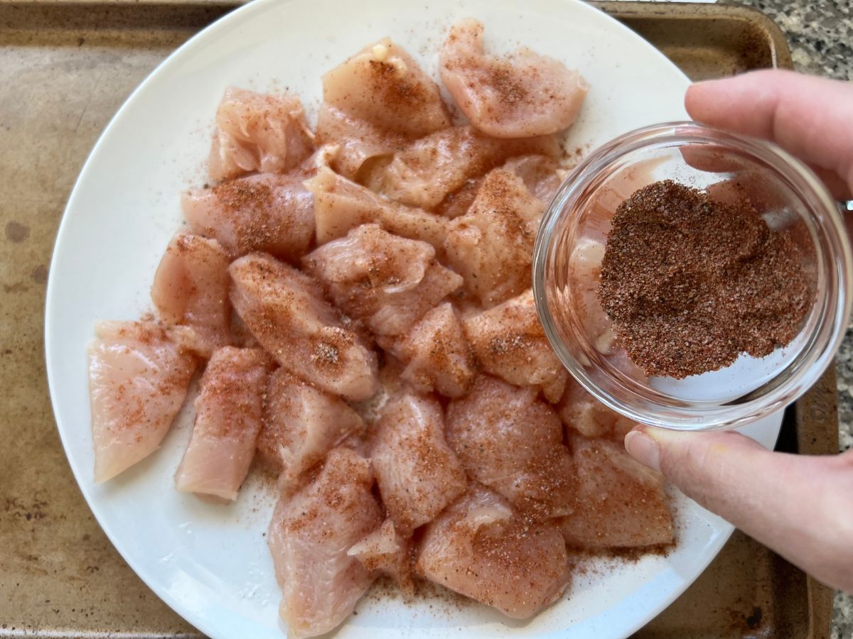 Homemade seasoning in a bowl being sprinkled over 1 inche chicken chunks.