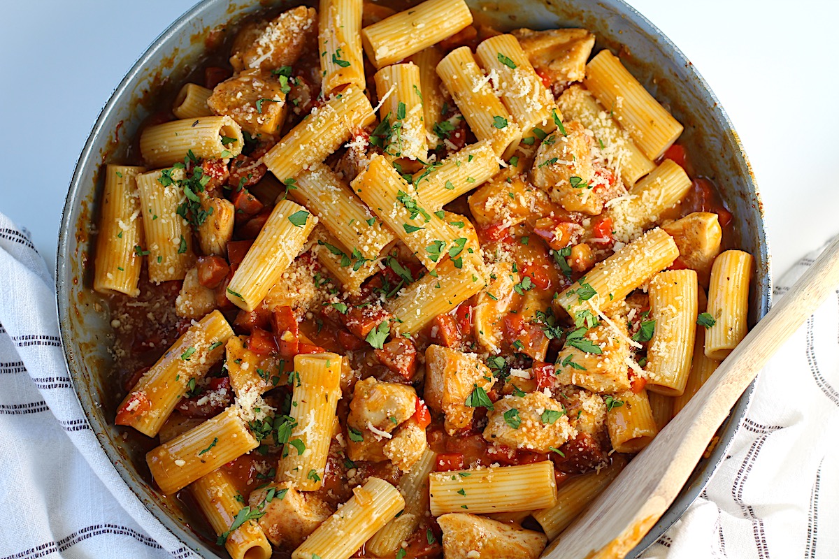 Top view of a pan filled with pasta with chorizo and chicken and sprinkled with chopped parsley and a wooden spoon resting on the edge.