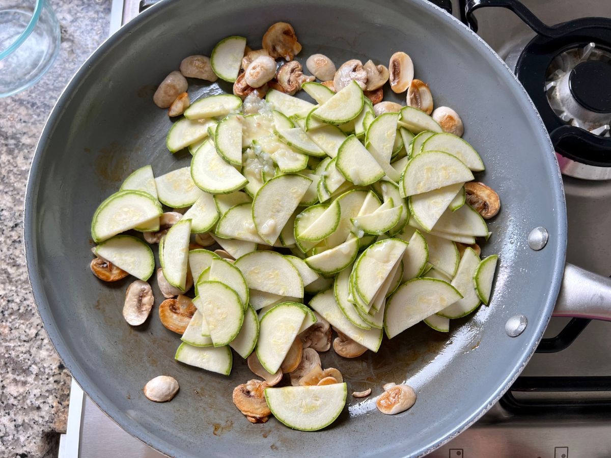 Garlic added over thinly sliced mushrooms and zucchini being sauteed in a skillet.