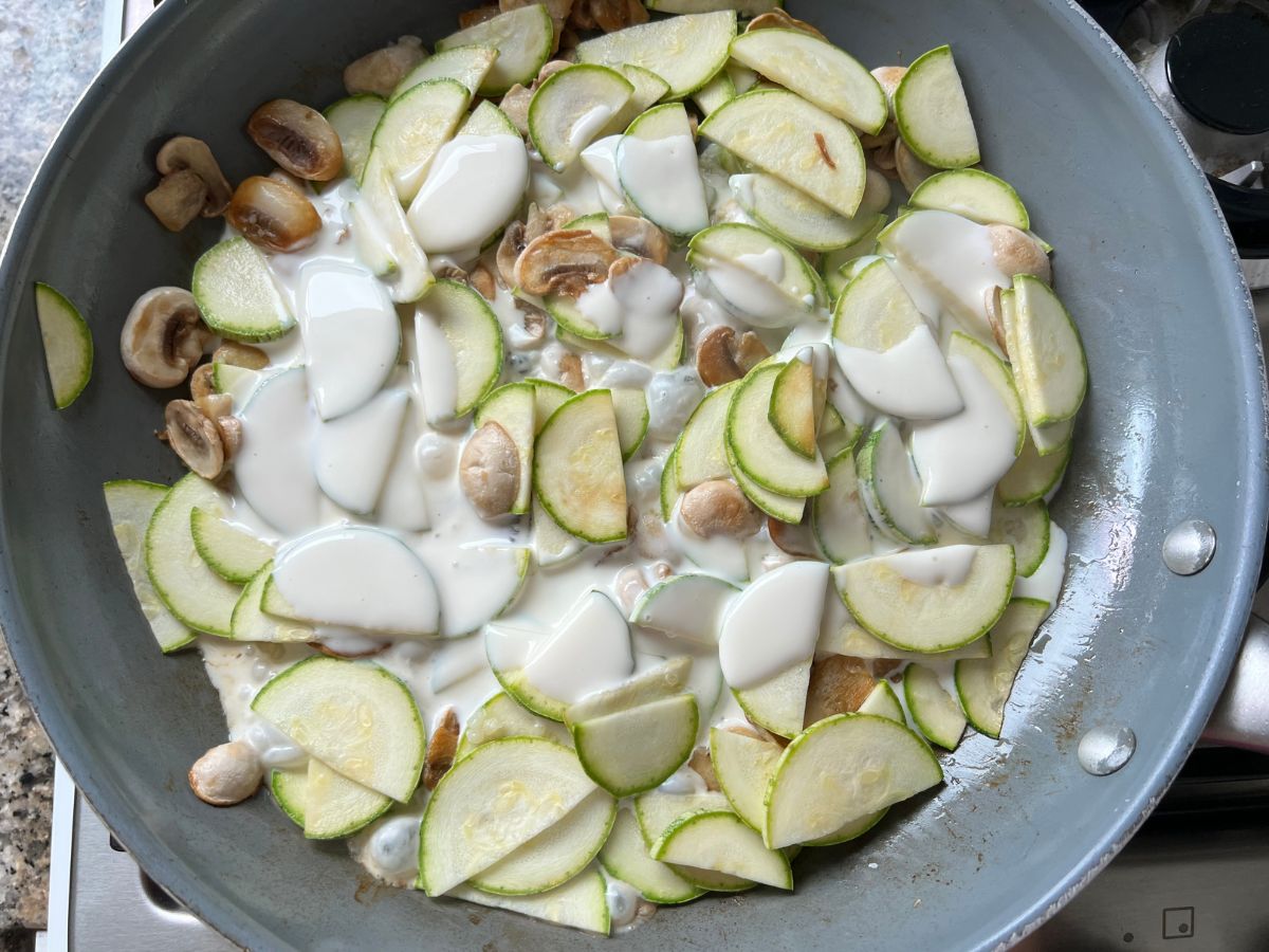 Heavy cream added to sauteed zucchini and mushrooms in a skillet.