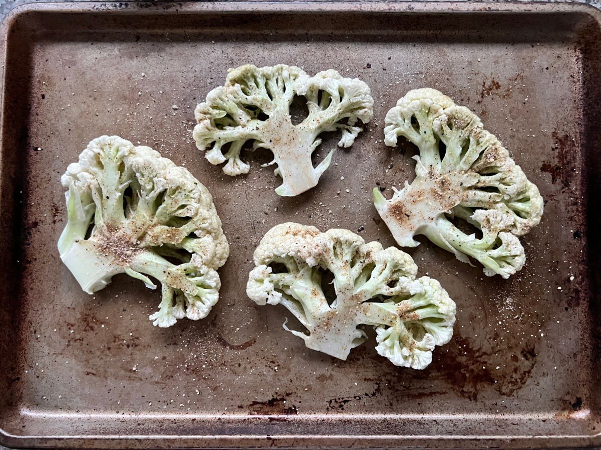 Seasoning sprinkled over raw cauliflower steaks on a baking sheet.