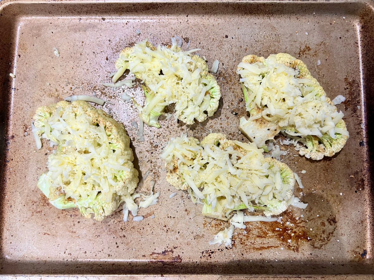 Shredded cheeses sprinkled over seasoned caulfilower steaks on a baking sheet, ready to roast or grill.