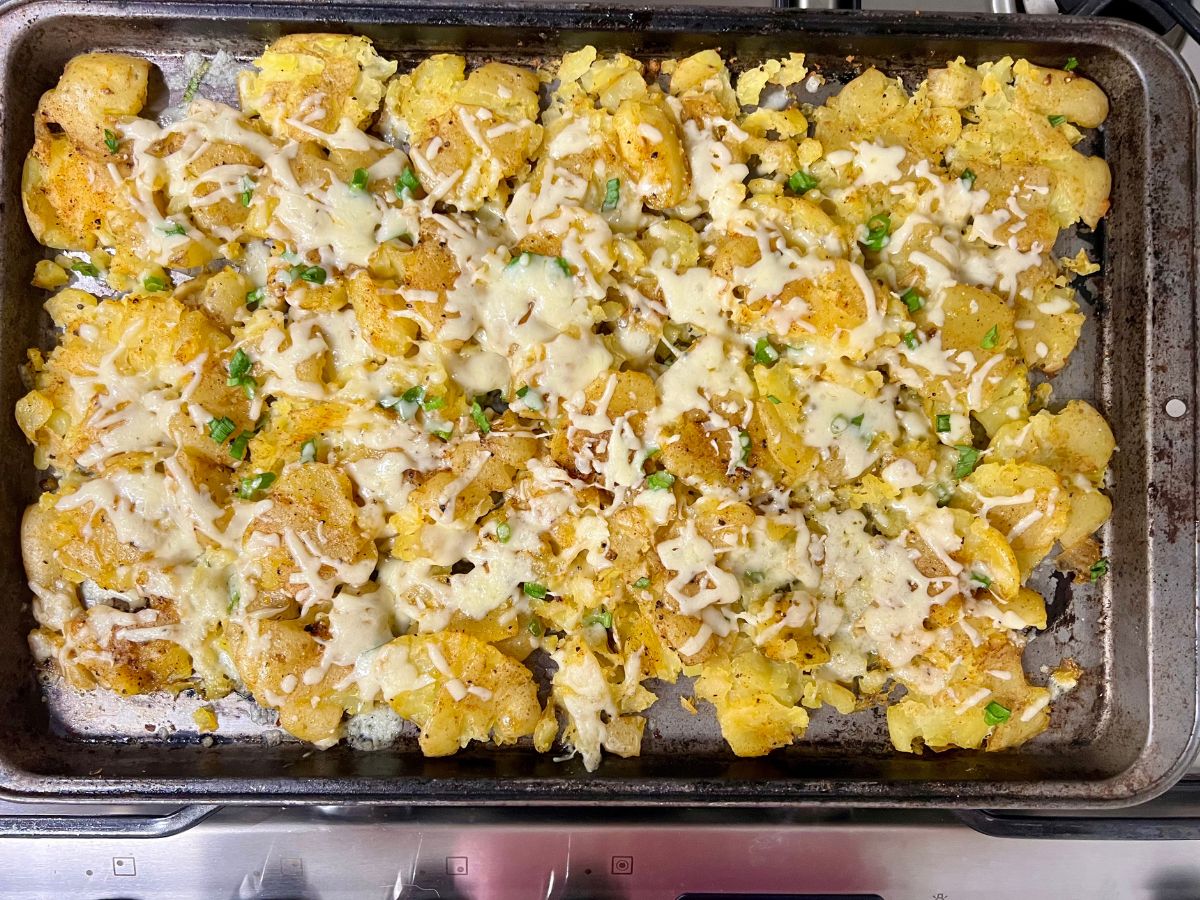 Melted cheese on top of seasoned smashed potatoes on a baking sheet with chopped chives sprinkled over.