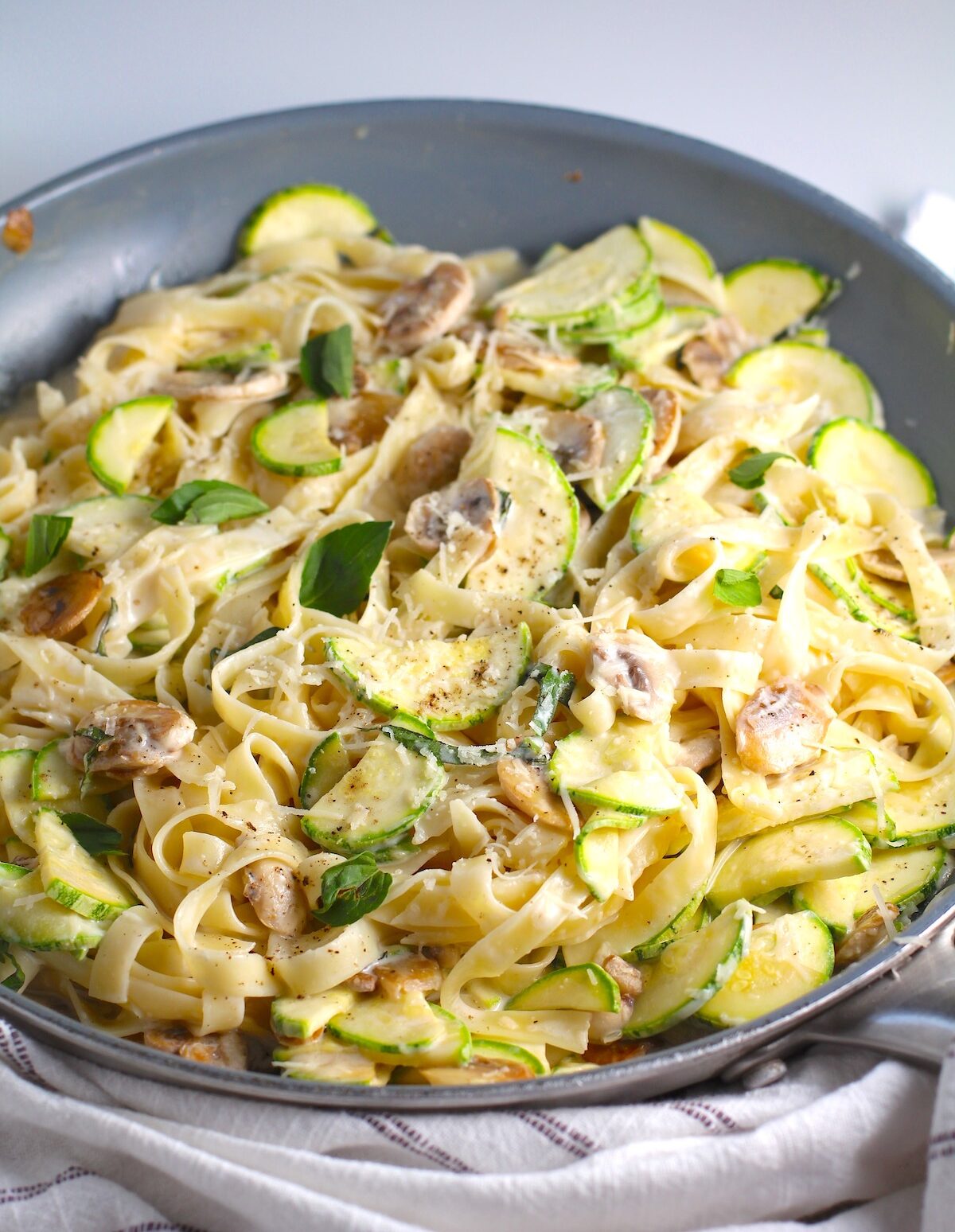 Creamy Fettuccine Pasta with Zucchini and Mushrooms in a skillet on counter with blue and white towel. Parmesan cheese is shredded on top.