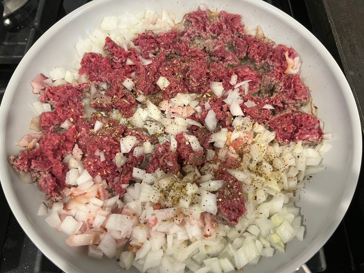 Ground beef, diced onions and chopped bacon in a skillet topped with seasonings.
