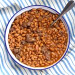 Bowl of Baked Beans with Ground Beef and Bacon with a spoon on top of a blue and white striped tablecloth.