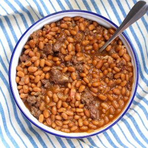 Bowl of Baked Beans with Ground Beef and Bacon with a spoon on top of a blue and white striped tablecloth.