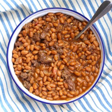 Bowl of Baked Beans with Ground Beef and Bacon with a spoon on top of a blue and white striped tablecloth.