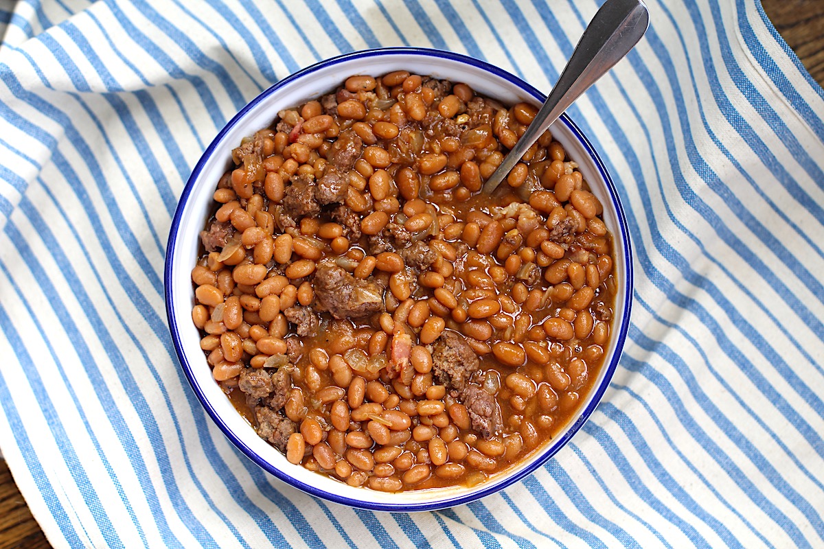 Bowl of Baked Beans with Ground Beef and Bacon with a spoon on top of a blue and white striped tablecloth.