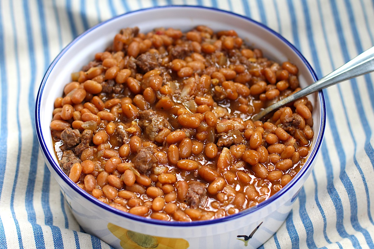 Bowl of Baked Beans with Ground Beef and Bacon with a spoon on top of a blue and white striped tablecloth.