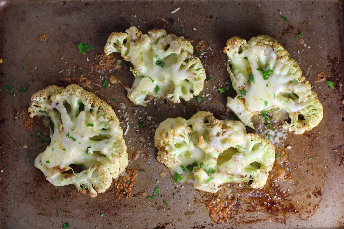 Four Cheesy Cauliflower steaks roasted on a sheet pan with cheese melted on top and edges browned and crispy.
