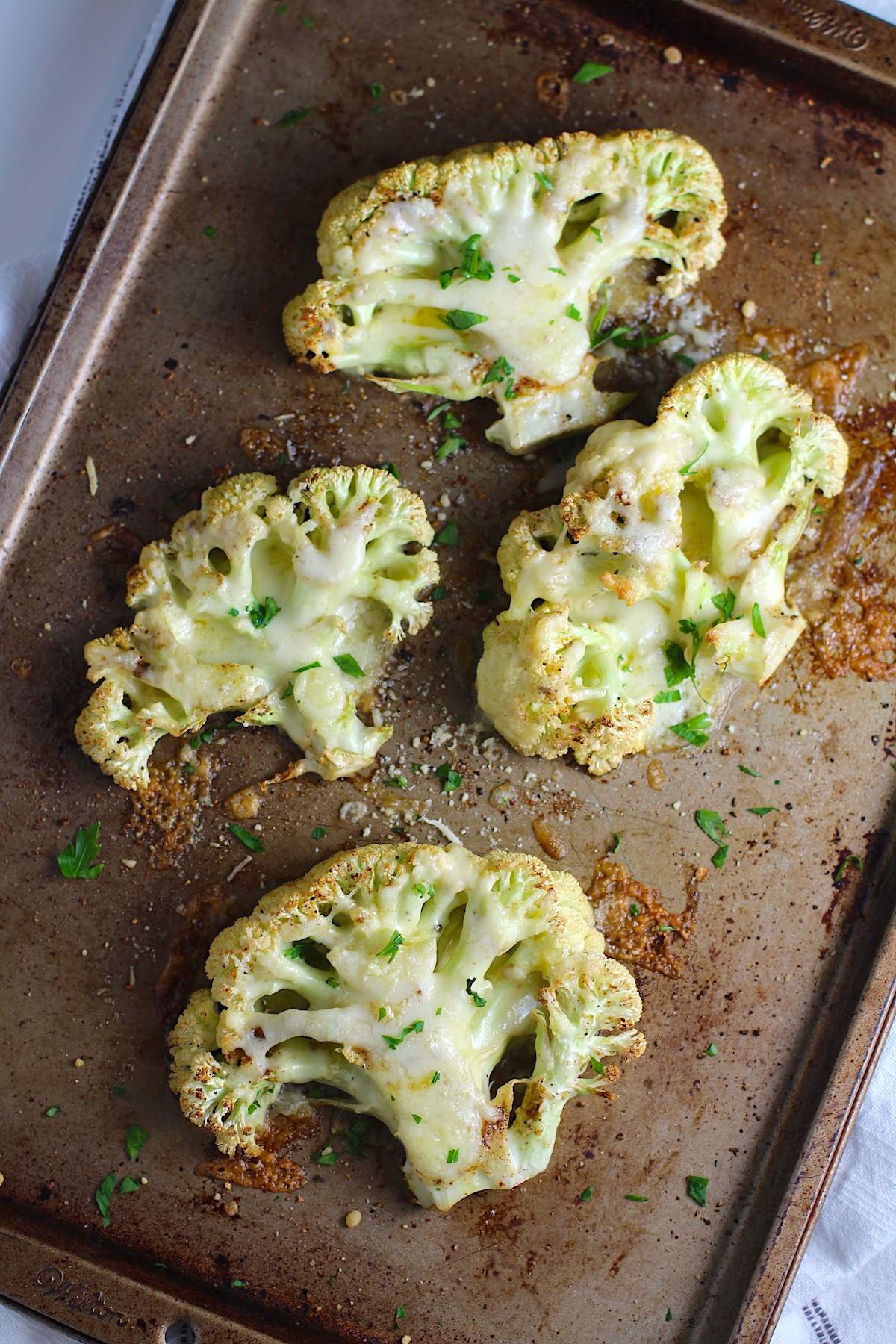 Four Cheesy Cauliflower steaks roasted on a sheet pan with cheese melted on top and edges browned and crispy.