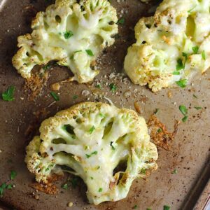 Four Cheesy Cauliflower steaks roasted on a sheet pan with cheese melted on top and edges browned and crispy.