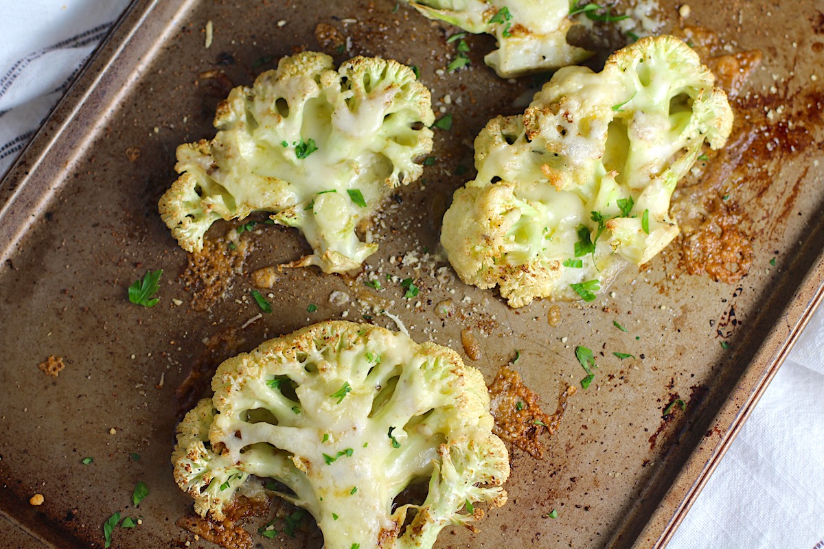 Four Cheesy Cauliflower steaks roasted on a sheet pan with cheese melted on top and edges browned and crispy.