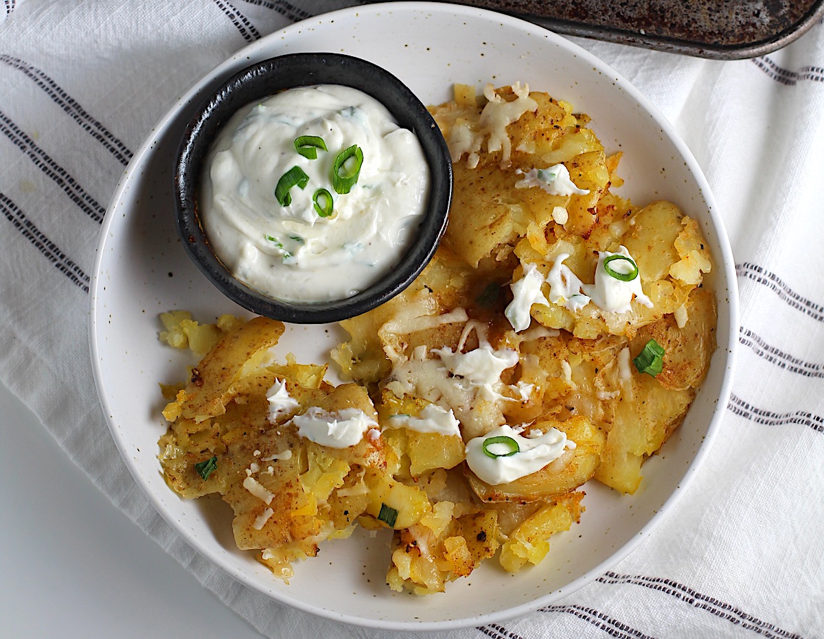 Crispy cheesy smashed potatoes recipe with smashed potatoes on a plate with dollops of sour cream and sliced scallions on top, along with a small bowl of sour cream.