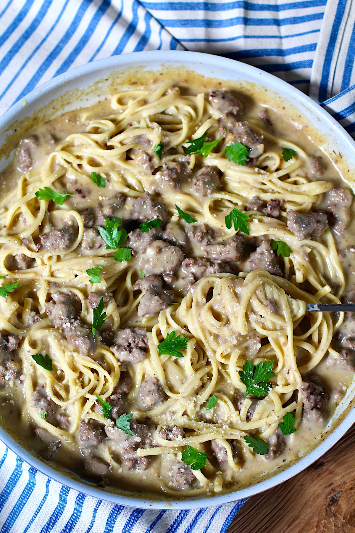 Creamy Alfredo Ground Beef Pasta with fettuccine pasta and parsley on top in a skillet on a wood table with blue and white towel.